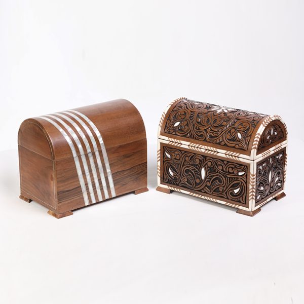 Two wooden chests: the left one is smooth with three silver stripes, while the right one is intricately carved with floral and leaf patterns accented in white. Both showcase different styles of craftsmanship.